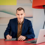 Mann im Sakko sitzt mit MacBook und Grafiktablett am roten Schreibtisch beim Business Fotoshooting München mit Natalia Schuchardt